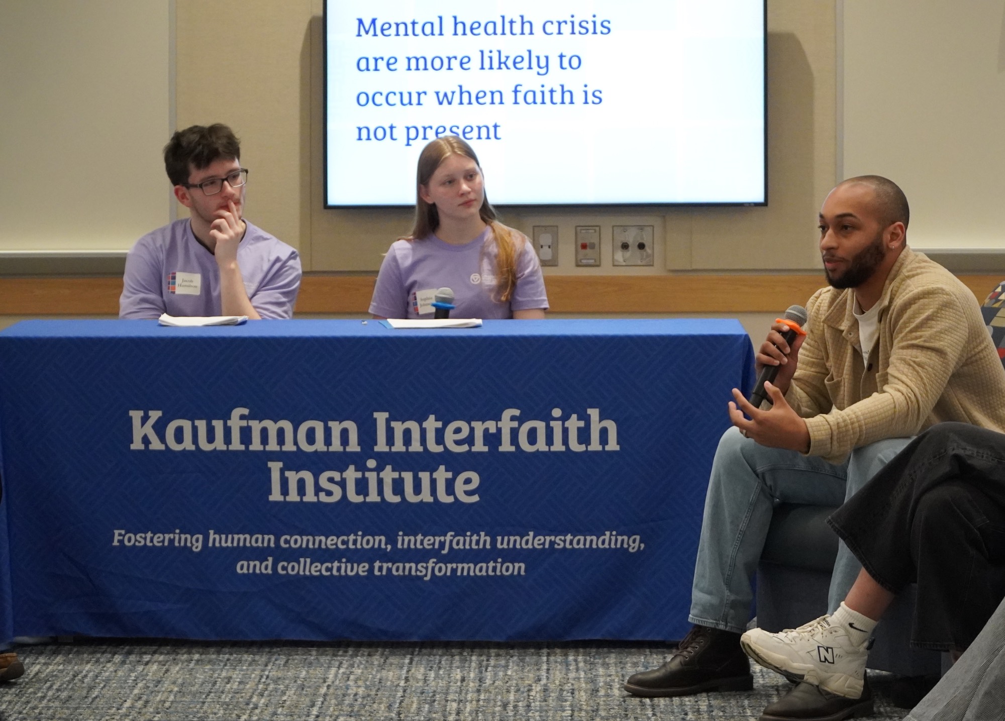 Two students sitting behind a table moderating a discussion with one person sitting in a chair speaking into a microphone.